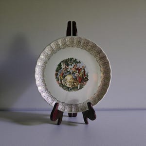 Peut inclure: Une assiette ronde en céramique à bord festonné ornée d'un motif en filigrane doré sur le pourtour. Le centre de l'assiette présente une illustration colorée de deux personnages en tenue d'époque dansant en extérieur sous des arbres verts.