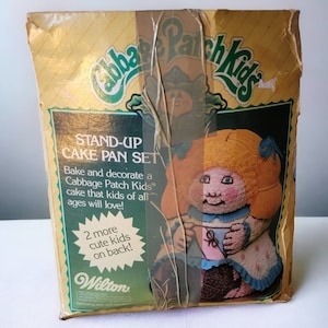 May include: A yellow and green box with the words "Cabbage Patch Kids" on it. The box contains a stand-up cake pan set. The front of the box features a Cabbage Patch Kid doll holding a pink cup. The text on the box reads "Stand-Up Cake Pan Set" and "Bake and decorate a Cabbage Patch Kids cake that kids of all ages will love!"