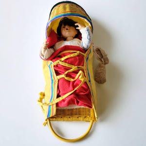 May include: A yellow and red doll cradle with a doll inside. The cradle is made of woven wicker and has a yellow fabric lining. The doll is wearing a white dress and has brown hair.