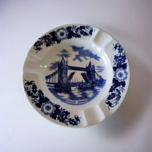 Może przedstawiać: Biała ceramiczna popielniczka z niebieską ilustracją Tower Bridge i kwiatowymi akcentami. Popielniczka ma okrągły kształt z czterema wgłębieniami na papierosy. Na popielniczce wydrukowano napis "Tower Bridge".