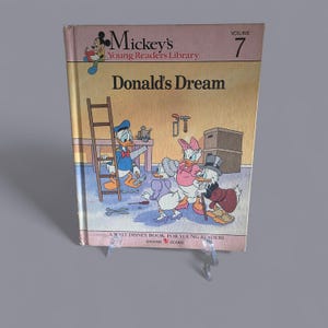 Puede incluir: Una copia vintage de "Donald's Dream", Volumen 7 de Mickey's Young Readers Library. La portada presenta a Donald Duck, Daisy Duck y Scrooge McDuck en una colorida ilustración. El libro es un libro de Walt Disney para jóvenes lectores, publicado por Bantam Books.