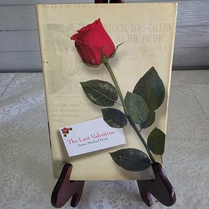 Puede incluir: Un libro de tapa dura titulado The Last Valentine de James Michael Pratt con una cubierta de color crema que presenta texto al estilo de un periódico antiguo y una rosa roja con hojas verdes colocada en diagonal sobre la portada.