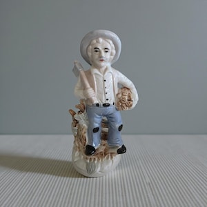 Puede incluir: Figura de cerámica de un hombre con camisa blanca, pantalones azules y sombrero, sosteniendo un hacha y un fardo de heno. La figura está sobre una base decorativa con diseño de tronco de árbol. La paleta de colores incluye blanco, azul y marrón.