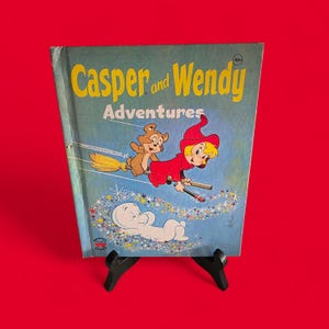 以下が含まれることがあります： ヴィンテージの「キャスパーとウェンディの冒険」の本。表紙には、おなじみの幽霊キャスパー、良い小さな魔女ウェンディ、そしてほうきに乗った茶色のクマが描かれています。タイトルは黄色で、背景は青、縁は赤です。