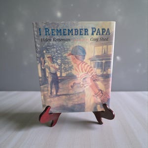 Może przedstawiać: Vintage książka zatytułowana "I Remember Papa" autorstwa Helen Ketteman, z ilustracjami Grega Sheda. Okładka przedstawia scenę baseballową z dwiema postaciami. Książka jest prezentowana na małym, czerwonym drewnianym stojaku.