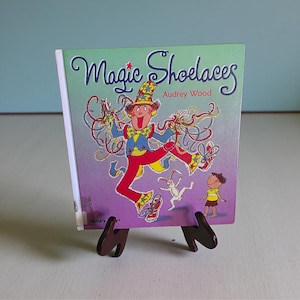 May include: A children's book titled "Magic Shoelaces" by Audrey Wood. The cover features a whimsical illustration of a clown-like figure with colorful shoelaces, a rabbit, and a child. The book is displayed on a small wooden easel.