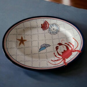 Op de afbeelding: Witte ovale serveerschaal met een blauwe en rode rand. De schaal heeft een nautisch thema met een zeester, schelpen en een krab.
