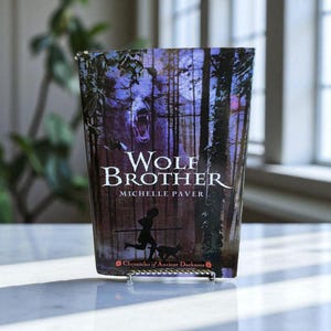 May include: A hardcover book titled "Wolf Brother" by Michelle Paver. The cover features a wolf's face, a silhouetted figure running with a dog, and forest imagery in shades of purple and brown. The book is propped up on a silver metal stand.