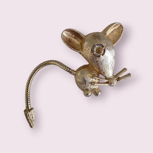 May include: A gold-toned mouse brooch with a textured finish. The mouse has large ears, a small nose, and a curved tail. A clear rhinestone is set in the center of the mouse's head. The brooch is a vintage accessory.