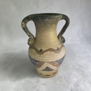 Puede incluir: Un jarrón de cerámica con dos asas, que presenta un color base beige y un acabado texturizado y desgastado. El cuerpo está decorado con patrones geométricos en tonos marrón, gris oscuro y verde apagado, dispuestos en bandas horizontales.