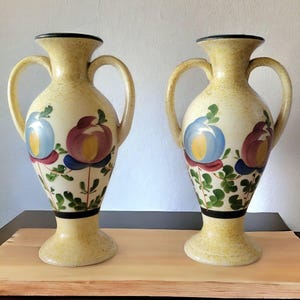 May include: Two ceramic vases with floral designs. The vases are yellow with blue, red, and green painted flowers. The vases have two handles and a narrow neck.
