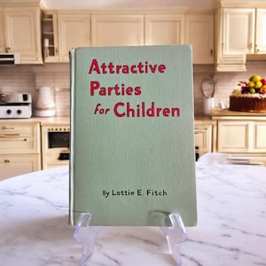 May include: A vintage book titled "Attractive Parties for Children" by Lottie E. Fitch. The book has a light green cover with red lettering and is displayed on a clear plastic stand. The background shows a kitchen setting.