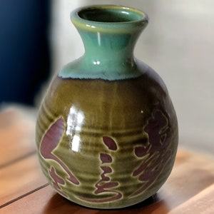 May include: A small, round, green and brown ceramic sake bottle with a wide mouth and a teal glaze. The bottle has a raised design of Japanese characters in a reddish-brown color.
