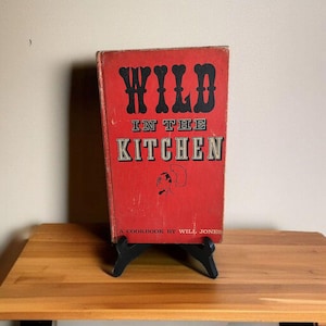 Puede incluir: Un libro de cocina vintage titulado "Wild in the Kitchen" con una portada roja y letras negras. El libro está apoyado en un soporte negro. La portada presenta el título, una ilustración de un chef y el nombre del autor.