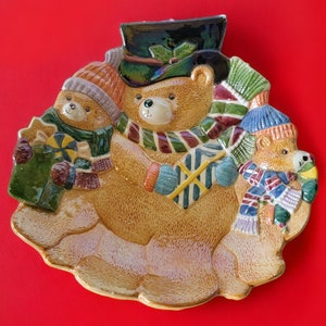 Puede incluir: Un plato de cerámica con tres osos de peluche marrones que llevan gorros y bufandas de invierno. Los osos sostienen regalos y están rodeados de un paisaje nevado.