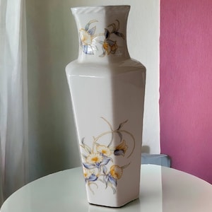 May include: A white ceramic vase with a geometric shape and floral design. The vase features yellow, blue, and green flowers with gold accents.