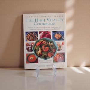 May include: A cookbook titled "The High Vitality Cookbook" by Maggie Pannell, displayed on a clear stand. The cover features a collage of food images, including a teal bowl of shrimp and vegetables. The book promises over 70 recipes for health, energy, and fitness.