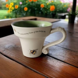 Op de afbeelding: Crèmekleurige keramische mok met een uitlopende vorm en een gebogen handvat. De mok heeft een bijenillustratie en de tekst "the welcoming hum of the honey bee". De binnenkant is lichtgroen. De mok staat op een houten tafel.