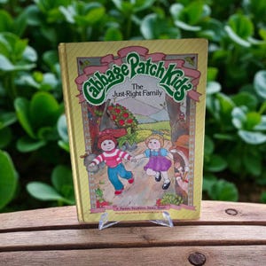 May include: A vintage children's book titled "Cabbage Patch Kids: The Just-Right Family." The cover features illustrations of two Cabbage Patch Kids running, with a forest scene in the background. The book is displayed on a clear acrylic stand.