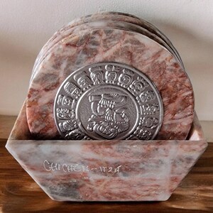 May include: Set of six pink and white marble coasters with a silver Mayan calendar design in the center. The coasters are in a matching marble holder with the inscription "Chichen Itza" on the front.