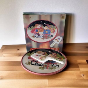 Op de afbeelding: Een keramische taartplateau en serverset met een winterse feestthema. De plaat heeft een rood en groen ontwerp met twee sneeuwpoppen die op een slee rijden. De server is wit met een sneeuwpopontwerp.