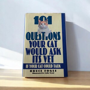 Puede incluir: Libro titulado "101 Preguntas que tu gato le haría a su veterinario" con una imagen de gato. El libro tiene un fondo crema con texto azul y un borde azul. Autor: Bruce Fogle.