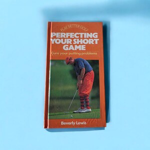 May include: An orange book titled "Perfecting Your Short Game" with a photo of a golfer putting. The book cover also includes the text "Play Better Golf" and "Cure your putting problems". The author's name, Beverly Lewis, is at the bottom.