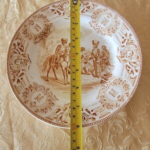 May include: A white ceramic plate with a brown design depicting a military scene. The plate is 9 inches in diameter and features the text "Bataille de Geneve" and the letter "N".