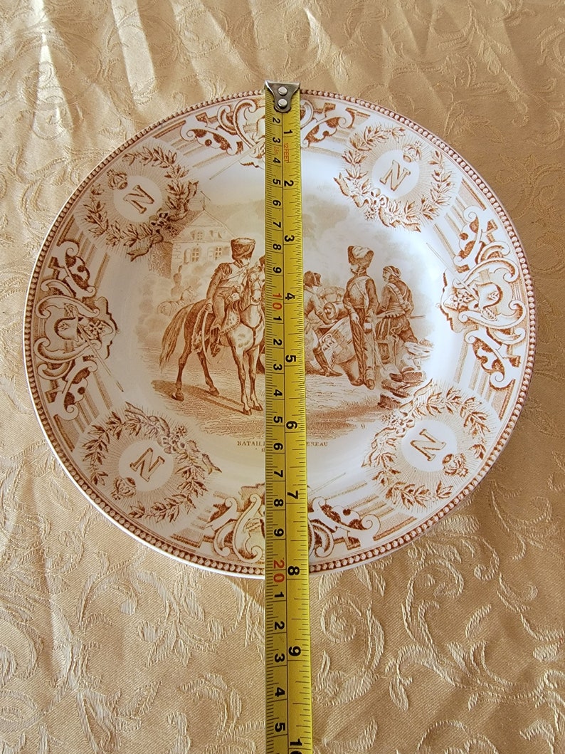 May include: A white ceramic plate with a brown design depicting a military scene. The plate is 9 inches in diameter and features the text "Bataille de Geneve" and the letter "N".