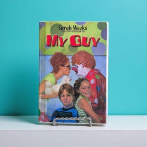May include: A hardcover book titled "My Guy" by Sarah Weeks. The cover art features a woman kissing a small white dog, a clown, and two children. The book is displayed on a silver metal book stand against a teal background.