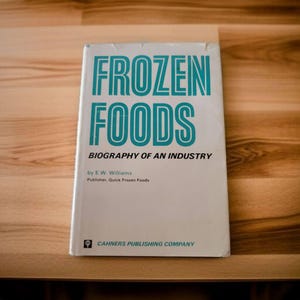 May include: A white book with the title "FROZEN FOODS" in blue text. The subtitle is "BIOGRAPHY OF AN INDUSTRY" in smaller blue text. The author is listed as "E.W. Williams" and the publisher is "Quick Frozen Foods". The book is published by "CAHNERS PUBLISHING COMPANY".