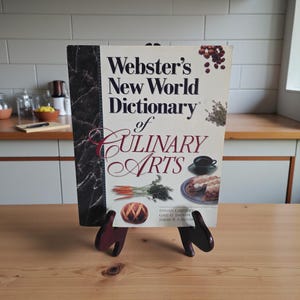 May include: A hardcover book titled "Webster's New World Dictionary of Culinary Arts" is displayed on a wooden stand. The book cover features illustrations of food items and the title in a decorative font. The stand is dark red and has a unique design.