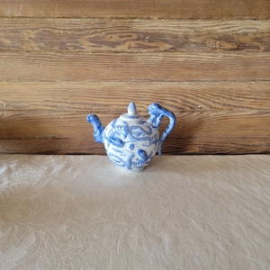 May include: A white teapot with a blue and white floral design. The teapot has a spout and a handle on either side. The teapot is sitting on a white tablecloth.