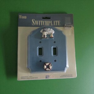 May include: A blue wooden switchplate with a nautical theme. The switchplate has two rectangular openings for switches and is decorated with a lighthouse, sailboat, and a life preserver. The text "WOOD" and "SWITCHPLATE" are visible.