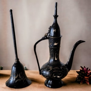 May include: Two black metal decorative items with intricate designs. The larger item is a teapot with a spout and a lid. The smaller item is a bell-shaped object with a long handle.
