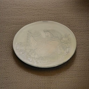 Puede incluir: Un plato redondo de cerámica blanquecina con un diseño en relieve detallado. El diseño representa una figura con barba y sombrero, posiblemente Papá Noel, con otros elementos festivos. El plato tiene un borde ligeramente elevado.