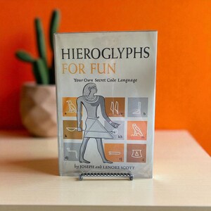 May include: A book titled "Hieroglyphs For Fun" with the subtitle "Your Own Secret Code Language." The cover features an illustration of an Egyptian figure and hieroglyphic symbols. The book is propped up on a silver metal stand. The background is orange.
