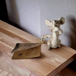May include: A ceramic mouse figurine with a tag attached by a gold string. The mouse is off-white with brown accents. The tag is brown with text. The figurine is on a wooden surface.