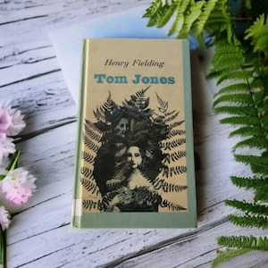 Puede incluir: Un libro de bolsillo vintage titulado "Tom Jones" de Henry Fielding. La portada presenta una ilustración en blanco y negro de una mujer con helechos.