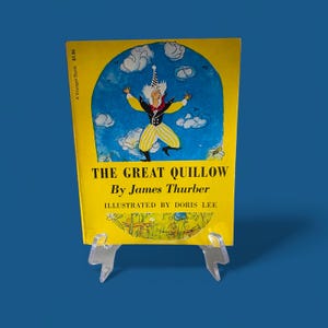 May include: A vintage copy of "The Great Quillow" by James Thurber, illustrated by Doris Lee, displayed on a clear stand. The book has a yellow cover with an illustration of a whimsical character against a blue sky with clouds. The book is a Voyager Book.