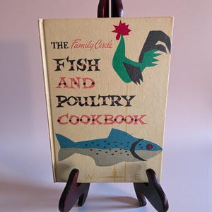 May include: A vintage cookbook titled "The Family Circle Fish and Poultry Cookbook" is displayed on a wooden easel. The cover features a rooster, a fish, and the title in black and red lettering. The book has a cream-coloured background.