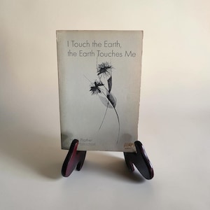 May include: A softcover book titled I Touch the Earth, the Earth Touches Me by Hugh Prather, featuring a black and white line drawing of two flowers on the cover. The book is displayed upright on a dark brown wooden stand.