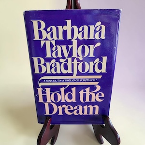 Op de afbeelding: Een hardcover boek van Barbara Taylor Bradford, getiteld "Hold the Dream". Het boek heeft een diepblauwe omslag met witte letters. De titel en de naam van de auteur zijn duidelijk zichtbaar. Het boek staat op een houten standaard.