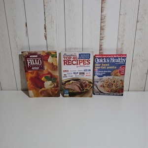 May include: Three cookbooks are displayed, featuring titles like "ATHENS AWARD WINNING FILLO RECIPES APOLLO", "America's TOP 40 RECIPES", and "Quick & Healthy Our best low-fat pasta". The books are on a white surface against a white wood-paneled background.
