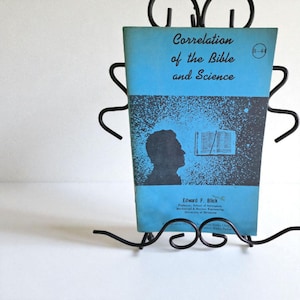 May include: A vintage book titled "Correlation of the Bible and Science" in a black metal book holder. The book has a light blue cover with black text and a silhouette of a man. The holder has a decorative, open design.