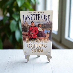 Peut inclure: Un livre intitulé "Beyond The Gathering Storm" de Janette Oke, présenté sur un présentoir en acrylique transparent. La couverture montre un homme en uniforme rouge et une femme en jupe violette, avec un paysage de montagne en arrière-plan.