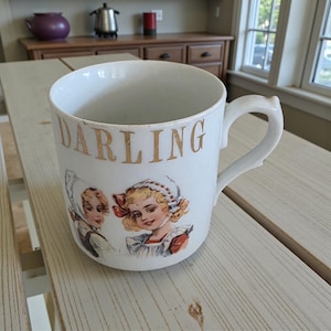 Puede incluir: Taza de cerámica blanca con la palabra "DARLING" en letras doradas. La taza presenta una ilustración vintage de dos niñas con gorros y vestidos. La taza tiene un asa curva.