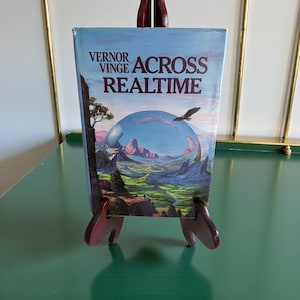 May include: A hardcover book titled "Across Realtime" by Vernor Vinge, displayed on a small wooden easel. The book cover features a landscape within a transparent sphere, with a flying bird. The book is set against a light blue background.