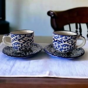 May include: Two blue and white patterned teacups with saucers on a white tablecloth. The cups have a blue and white speckled design.