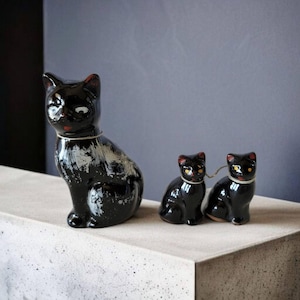 Puede incluir: Tres figuritas de gato de cerámica negra. El gato más grande está sentado con la cola enroscada alrededor de sus patas. Los dos gatos más pequeños están sentados uno al lado del otro, mirando hacia adelante.